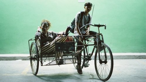 Filmstill of the opening film ASOG. Eine Person auf einem Fahrrad, eine weitere sitzt auf einem Beiwagen, der an dem Rad angebracht ist. Beide schauen in die Kamera